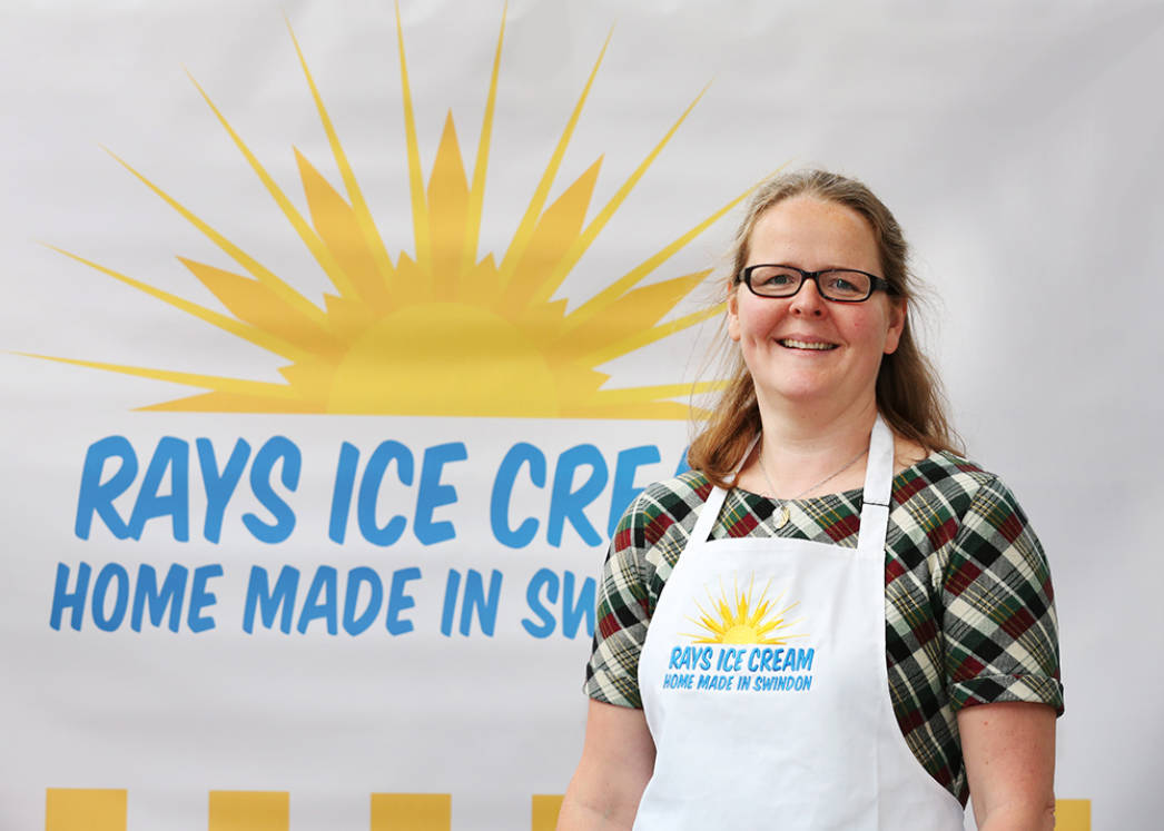 Hadi Brooks, founder of Rays Ice Cream pictured wearing a Rays branded apron and standing in front of a Rays Ice Cream logo